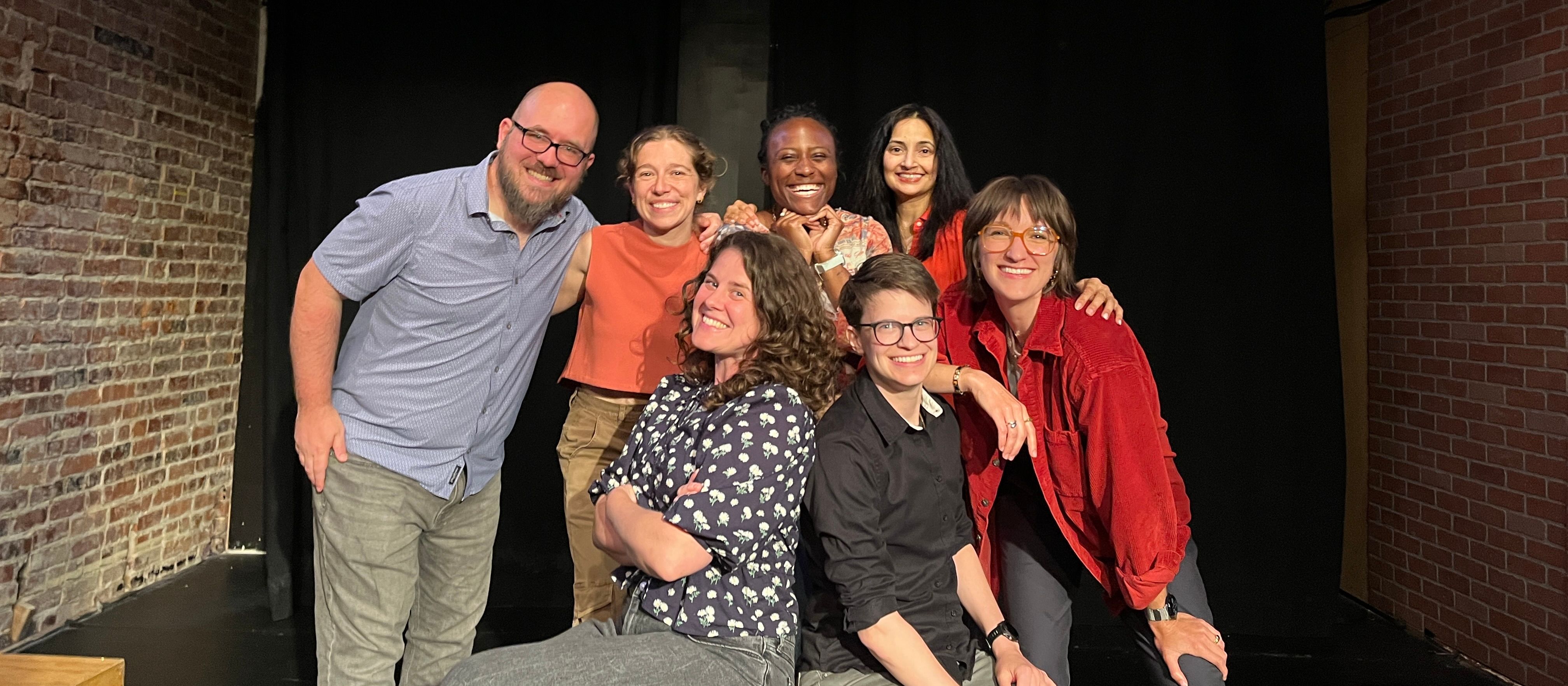 Seven Old AF cast members smiling and hugging on a dark stage
