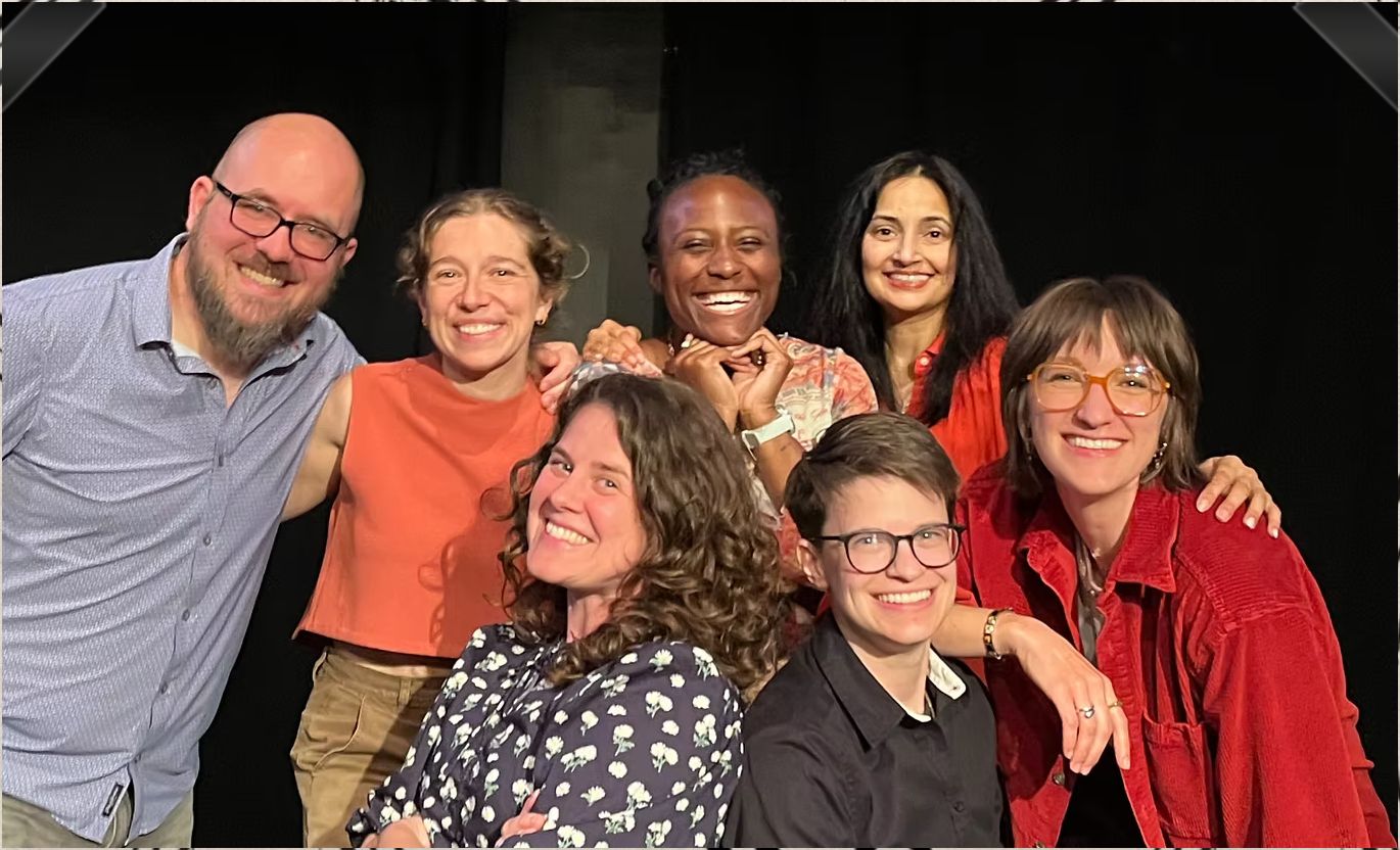 Seven Old AF cast members smiling and hugging on a dark stage
